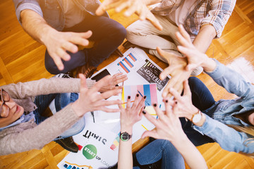 Young focused and motivated business people sitting on the floor of the office in the circle and raising arms after holding hands together in middle. Teamwork and togetherness concept.