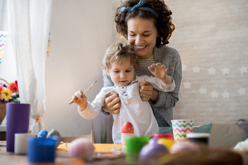 Fototapeta premium Portrait of happy young mother helping little toddler paint Easter eggs while making preparations for holiday at home