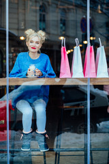 Vertical full body of tired woman with two bunch hairstyle and red lips smiling and drinking coffee after shopping at cafe in sunny morning. Photo taken trough the window.