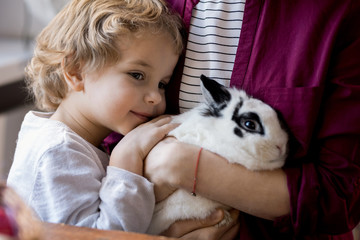 Fototapeta na wymiar Close up portrait of cute blond toddler hugging pet bunny and clinging to mom