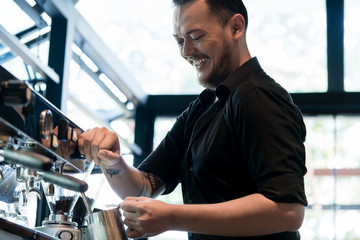 Low angle view of a young cheerful barista wearing black shirt while preparing coffee at an automatic machine in a modern coffee shop