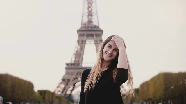 Young happy woman standing near the Eiffel tower in Paris, France, smiling at camera and sending the blow kiss.