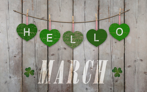 Hello March written on hanging green hearts and weathered wooden background, with clover