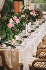 A beautiful table with a white tablecloth, plates for a dinner party, decorated with leaves of a palm tree, white candles and pink flowers with wicker chairs