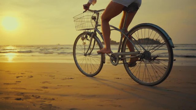 Woman in white and hat mooving on her bicycle along beach sand at summer time under burning sunset sky, lifestyle carefree.