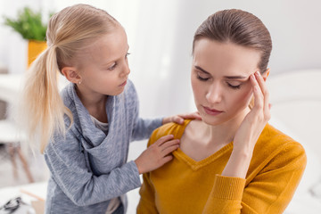 Do not upset. Sweet cute blond girl placing hand on moms shoulder while worrying about her and mother touching temple