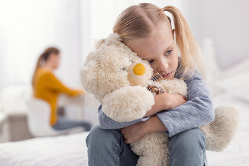 Always alone. Adorable appealing upset girl hugging plush bear while looking aside and posing on the blurred