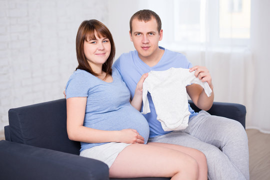 young beautiful pregnant couple sitting on sofa in living room and holding baby bodysuit