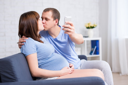 happy pregnant couple kissing and taking selfie photo with smart phone in living room