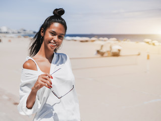 Portrait of smiling appealing Brazilian girl standing on the sunny summer beach and holding her sunglasses; with ocean horizon, sand and sunshades in defocused background; copy space for text message