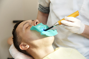 A young man with closed eyes in the dental chair. The dentist works with a dental polymerization lamp in the patient's mouth.