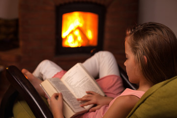 Young girl sitting in front of fireplace reading a book in the evening
