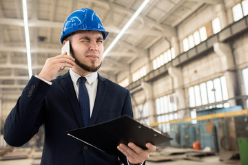 Bearded young entrepreneur wearing hardhat and classical suit talking to his business partner on mobile phone while standing at spacious production department of modern plant, portrait shot
