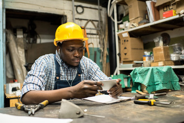 Concentrated African American lathe operator sitting at work bench and watching educational video on smartphone, interior of production department on background