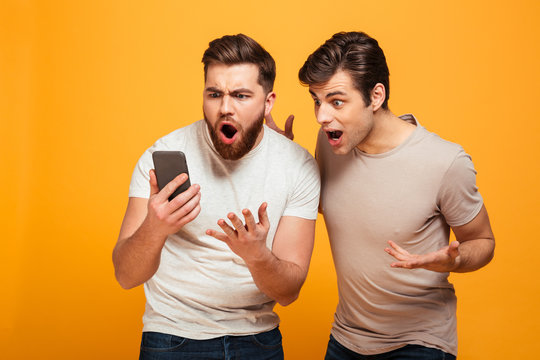 Photo of two men football fans emotionally gesturing while watching game on smartphone, isolated over yellow background