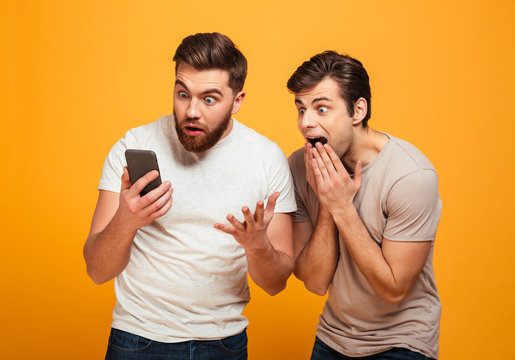 Photo of two men friends emotionally watching football game on cell phone and rooting for favorite team, isolated over yellow background