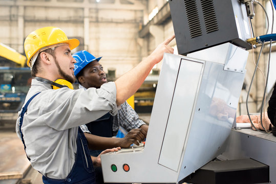 Multi-ethnic team of machine operators wearing hardhats and overalls discussing working process while standing in front of CNC, interior of production department on background