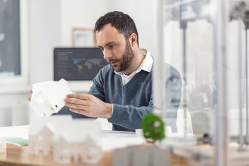 Flawless result. Pleasant handsome young engineer sitting at the table in the office and checking a 3D model of the house for any flaws and finding none - obrazy, fototapety, plakaty