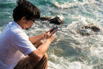 Asian man using phone typing message to chatting in social network while sitting on rock on sea.