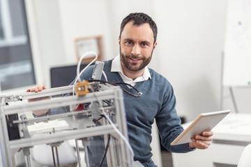 Futuristic technologies. Handsome bearded young man posing for the camera while controlling a 3D printer through a tablet in his hand while resting his hand on a printer