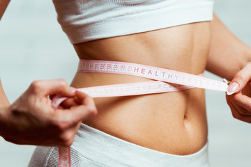 Beautiful, fit, young woman measuring her waist with a measuring tape, with word HEALHTY written on it, close up