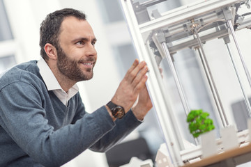 Interesting process. Pleasant young man resting his hands on the body of a 3D printer and watching closely the work of a 3D printer, controlling its performance