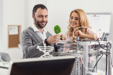 Satisfied engineers. Pleasant upbeat colleagues standing near a 3D printer and looking at a miniature tree model with smiles, being content with the result