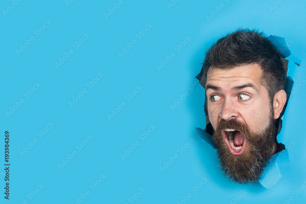 Wall mural View of male face through hole in blue paper. Surprised bearded man making hole in paper. Cute attractive man looking through hole. Copy space for advertising, to insert text or slogan. Discount, sale