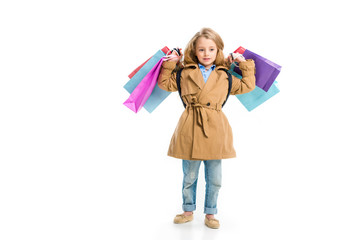 Kid in trench coat standing with different shopping bags in hands isolated on white