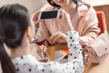 So shiny. Nice pleasant young woman holding her smartphone while taking a photo of her friends engagement ring