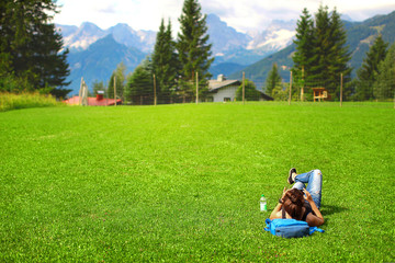 young woman tourist with a backpack and glasses resting on green grass and admiring the alpine mountains and meadows