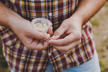 Close up cropped fisherman in checkered shirt holds in hands small white box with maggots, put bait on hook to fish with fishing rod. Man hold worms for fishing. Lifestyle, recreation, leisure concept