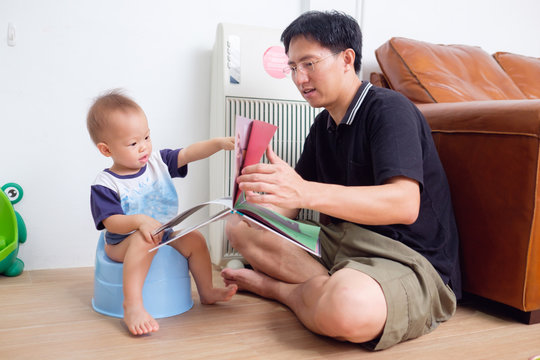 Father Training His Son To Use Potty, Cute Little Asian 1 Year Old / 18 Months Toddler Baby Boy Child Sitting On Potty And Reading Book With Young Dad In Living Room At Home. Potty Training Concept.