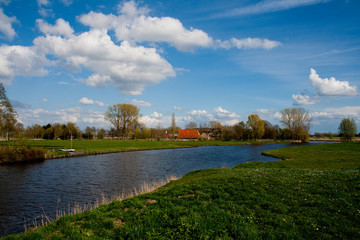 Landschaft in der Wesermarsch mit einem Siel, Weiden und im Hintergrund einem traditionellen Bauernhaus bei schönem Wetter, blauem Himmel mit weißen Wolken