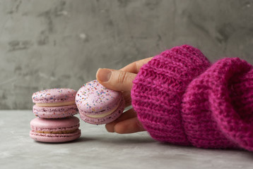 Hand of a girl holding a pink cake macaron or macaroon on grey background. Pink jumper.