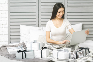 Smiling happy beautiful woman with gift boxes lying in bed with laptop. Morning surprise. Packing gifts.