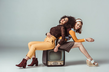 happy multicultural retro styled girls posing on vintage television on grey