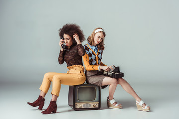 multicultural retro styled girls sitting on old tv and talking by stationary telephone