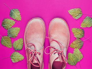 A pair of new brown leather men's shoes are on a pink background. Nearby there are green leaves. Trend background for a shoe store, fashion blog, travel agency. View from above