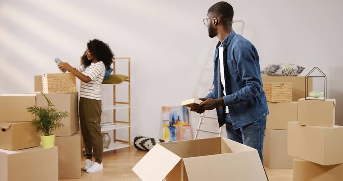 Handsome young African American man unpacking the carton box and showing a photo in the frame to his pretty curly girlfriend who standing behind. Couple moving in the new apartment. Indoor