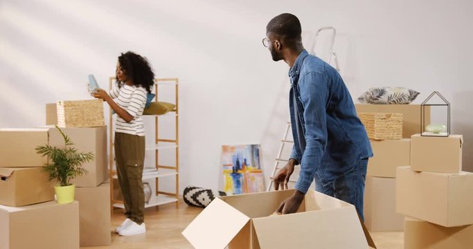 Handsome young African American man unpacking the carton box and his beautiful girlfriend coming closer from behind, taking a picture and looking for the place on the shelf while they moving in the