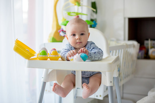 Cute little toddler boy, playing with plastic eggs, sitting in a white chair in a sunny living room