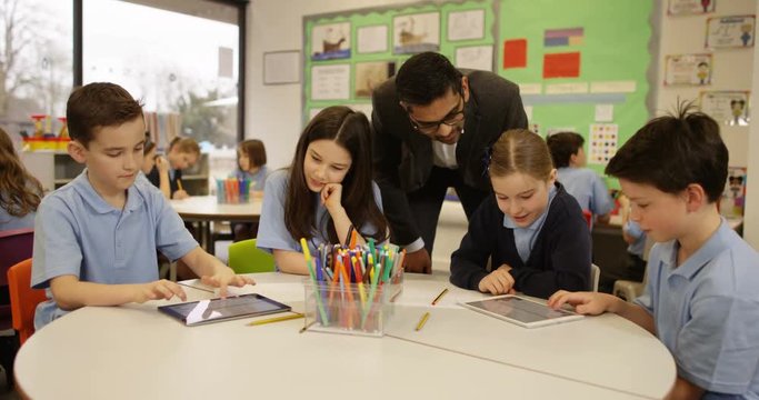 4K Happy young school children working on digital tablets in classroom with teacher helping out. Slow motion