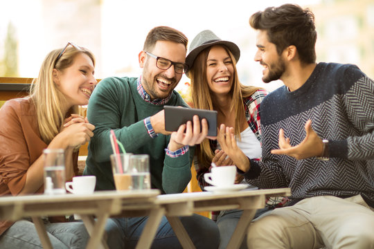 Group of four friends having a coffee together. Two women and two men at cafe talking laughing and enjoying their time using digital tablet.