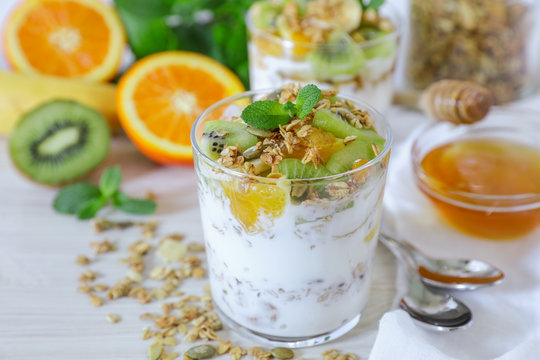 Tasty and healthy breakfast or snack: yogurt, granola and kiwi, orange, banana slices, honey, mint leaves on a white wooden background.