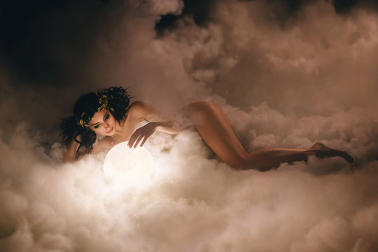 Fantasy woman attractive goddess, lies in  clouds and hugs moon. Creative whimsical hairstyle with unusual decoration golden tiara crown Greek style. Sexy Girl beautiful body. Artistic Photography