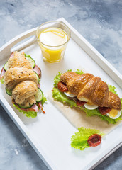 Croissant sandwich with tuna, hard boiled egg, salad and sun-dried tomatoes on white tray. Glass of orange juice.