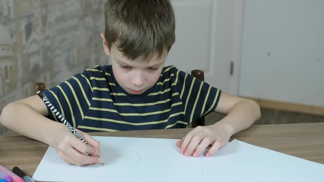 Boy of seven years in a striped t-shirt draws ovals with a simple pencil sitting at the table.