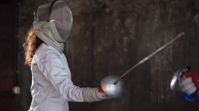 Woman with fencing mask on her face is training in a hall. She is attacking with epee and protecting, close-up during practice session of fencers school