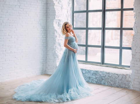 Young, pregnant woman posing in a luxurious, lush, blue dress with a long fairy train. The background is a light interior in Scandinavian style and a huge window. Art photo